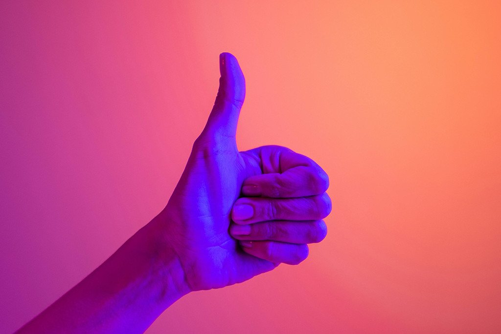 Een hand die een duim omhoog geeft, verlicht met blauwe en roze gradiëntverlichting op een zacht oranje en roze achtergrond, die doet denken aan positieve Google-recensies.