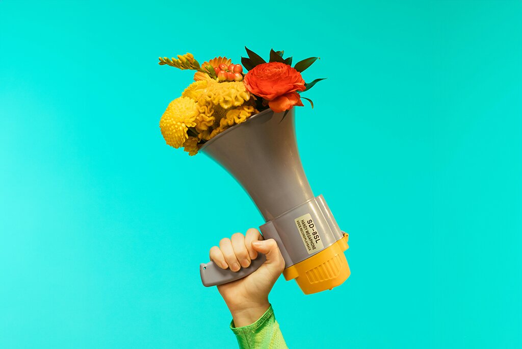 Een hand die een megafoon vasthoudt gevuld met een kleurrijk boeket bloemen tegen een blauwgroen achtergrond symboliseert de levendige essentie van inbound marketing.