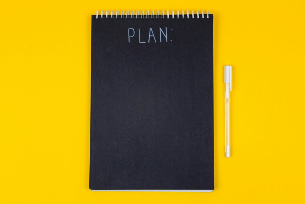 Zwart notitieboek met "PLAN" erop geschreven, gecombineerd met een witte pen, perfect om je WooCommerce checklist op te schrijven. Alles tegen een levendige gele achtergrond, het inspireert creativiteit en organisatie.